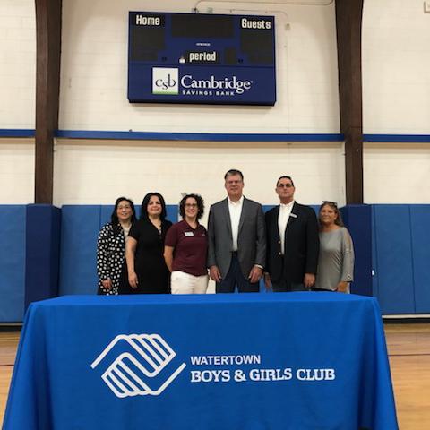 6 representatives from CSB or the Watertown Boys and Girls Club posing in a gym in front of a CSB digital scoreboard behind a table with the Watertown Boys & Girls Club logo on the tablecloth